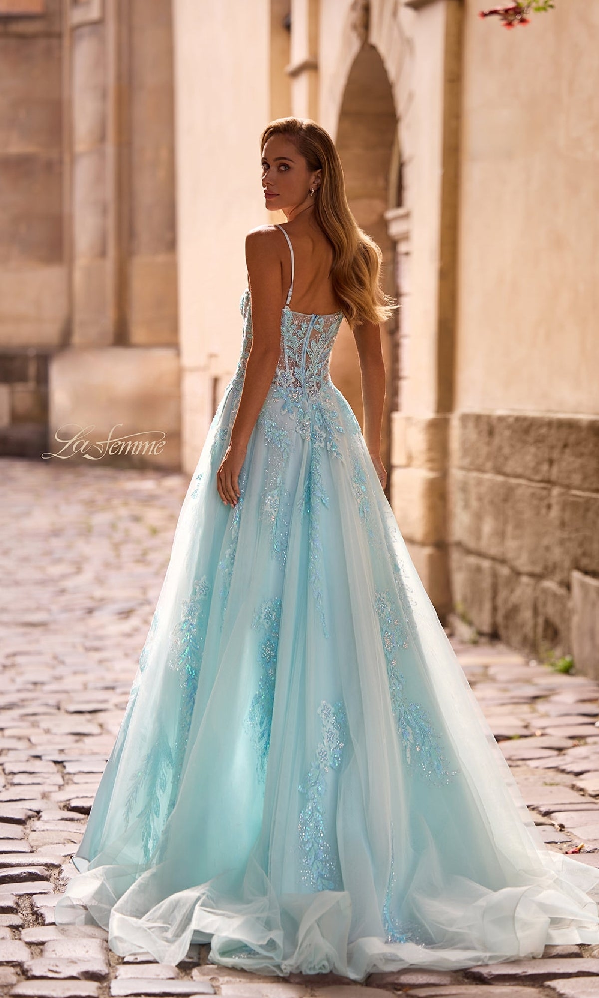 A woman in the La Femme 32810 long prom dress, featuring light blue tulle with beaded lace appliqué and a corset bodice, stands on a cobblestone street looking over her shoulder.